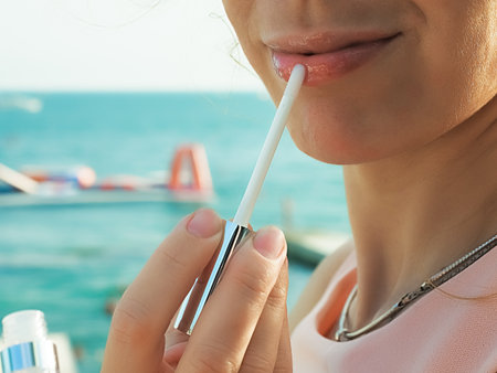 A woman paints her lips with lip gloss on a summer day against the backdrop of the sea. Close-up photo. Soft focus.の写真素材