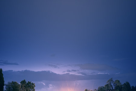 Evening blue sky with lightning sparkling in the clouds above the trees illuminated by the rays from...