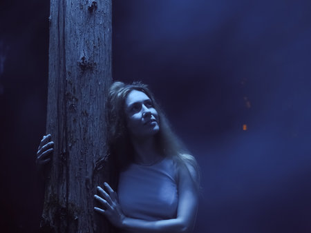 A smiling lady stands hugging a tree trunk in the blue illumination of a mystical night haze. Blurred image and noiseの写真素材