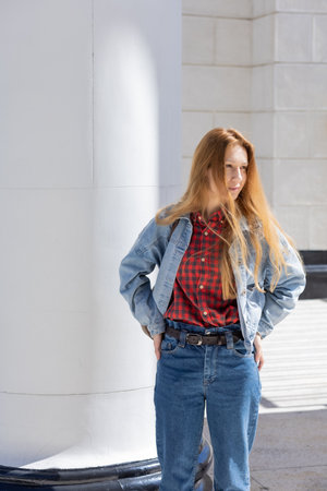 A woman in a casual denim outfit is standing near a wide white column illuminated by sunlight....