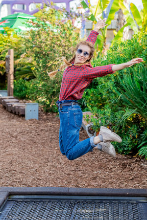 Joyful woman with pigtails jumping on a trampoline in a green southern amusement park on a spring day. Cheerful weekend. Finally a vacationの写真素材