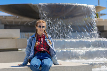 A woman in a plaid shirt and denim outfit sits on a curb at a cascading city fountain on a sunny spring day. Youth fashion. City walk.の写真素材