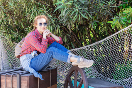 Russia, Sochi 14.05.2022. A smiling woman in a plaid shirt and jeans sits on a wooden pirate box near a green bush fenced with a rope fishing net. Nautical themed decor in an amusement park.のeditorial素材