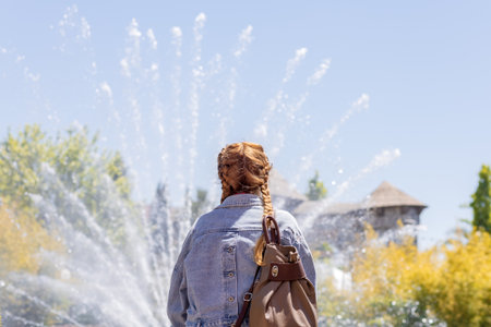 A woman with pigtails looks at a fan-shaped fountain splashing against the sky in a park on a sunny dayの写真素材