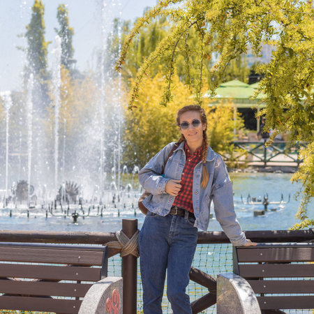 Joyful woman stands near lake with fountain under spring trees in sunny amusement parkの写真素材