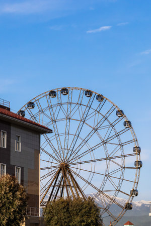 Russia, Sochi 14.05.2022. A tall ferris wheel behind a building and trees against the backdrop of snowy mountains and a bright blue skyのeditorial素材