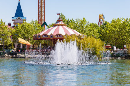 Russia, Sochi 14.05.2022. A fountain spouting from pond against the backdrop of multicolored bright carousels and spring green trees in an amusement parkのeditorial素材