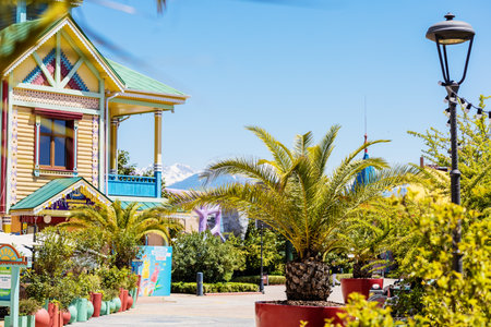 Russia, Sochi 14.05.2022. Exotic palm trees and a fabulous multi-colored wooden house against the backdrop of snowy mountains in a southern amusement park on a sunny spring dayのeditorial素材