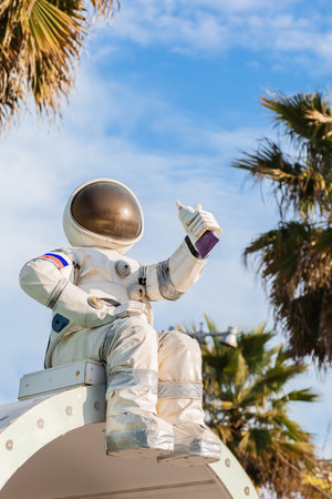 A cute figurine of an astronaut in a spacesuit holding a tube of food in his hand sits against a blue sky among palm trees in a south parkの写真素材