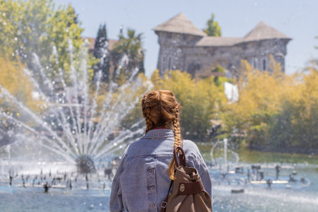 A woman with pigtails looks at the fountain gushing from the lake against the backdrop of trees and unusual park architecture. Selective focus.の写真素材