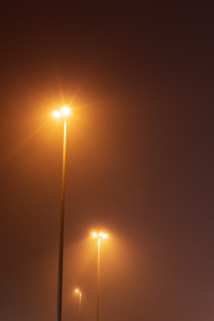 Street lights glow in the night foggy sky. Tall street lamps glow in the foggy night sky. Blurred...