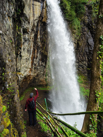 Tourist photographing a huge waterfall. Under the waterfall. Stand under a rock looking at a waterfall. Journey to the mountain waterfallの写真素材