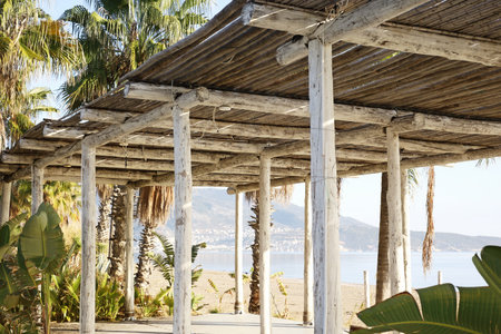 A rustic bamboo pergola provides shade along a beach with a clear view of mountains in the background and tropical plants surrounding the area.の写真素材