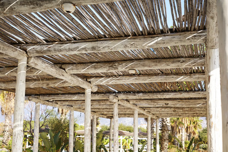 A rustic wooden pergola with thick beams forming the roof, casting shadows on the floor. Sunlight filters through the slats, highlighting the natural textures of the wood.の写真素材
