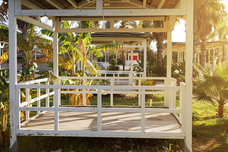 A white gazebo surrounded by banana plants in a lush garden. The structure shows signs of wear, and the garden is bathed in sunlightの写真素材