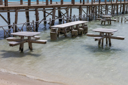 Serene image of a seaside pier featuring rustic wooden tables partially submerged in clear, gentle water. the tranquil setting exudes a peaceful ambiance, ideal for beachside gatherings or relaxation. the scene is enhanced by a sandy beach and a bright, inviting atmosphere, evoking the essence of a summer getaway. perfect for travel promotions, environmental themes, and coastal designs.の写真素材