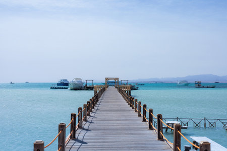 This tranquil image captures a wooden pier stretching into the turquoise water, with boats gently floating nearby under a clear blue sky. the peaceful scene evokes a sense of relaxation, making it ideal for travel promotions or vacation brochures. the calm ocean and bright horizon emphasize a serene maritime setting, perfect for those dreaming of a summer getaway.の写真素材