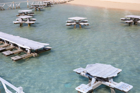 A serene scene featuring weathered wooden picnic tables floating on turquoise waters, creating a unique and tranquil beach setting. the clear blue water and sandy backdrop enhance the calm, summery atmosphere, perfect for relaxation or a picturesque vacation spot. ideal for travel content, depicting leisure and natural beauty.の写真素材