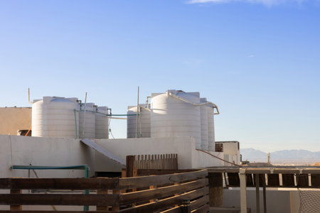 The image captures a series of white water tanks on a rooftop under a clear blue sky, with a distant mountain range in the background. the tanks, part of an urban infrastructure, highlight themes of sustainability and resource management. sunlight casts soft shadows, adding depth and contrast to the scene. this is suitable for illustrating concepts related to residential architecture or environmental engineering.の写真素材