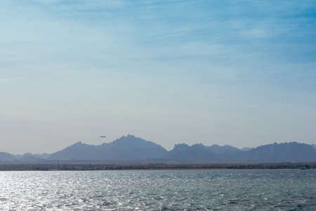 Calm and expansive seascape with gentle waves reflecting the soft sunlight, set against a backdrop of distant mountains. a small airplane adds a sense of journey to the wide-open sky. the scene conveys tranquility and the spirit of adventure, making it ideal for travel-themed materials or serene landscape settings. the pale blue hues and vast horizon evoke peace and exploration.の写真素材