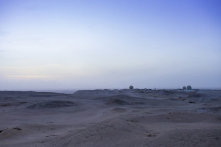 Expansive desert scene at dusk, featuring a vast horizon and remote structures under a calming twilight sky. earth tones dominate the barren landscape, with subtle gradients in the atmosphere creating a serene and isolated mood. ideal for themes of exploration, solitude, and natural beauty.の写真素材