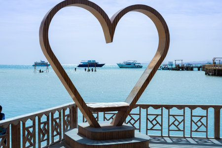 Charming scene showcasing a large wooden heart sculpture overlooking azure waters with boats in the distance. the bright blue sky and calm sea create a peaceful ambiance, perfect for romantic getaways or travel-themed projects. this image is the essence of love and leisure by the ocean, making it ideal for vacation promotions and serene designs.の写真素材