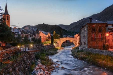A village's lights illuminate the river & the buildings at the evening time in a beautiful scene with a great variety of colors at Esterri d'Aneu, Spainのeditorial素材