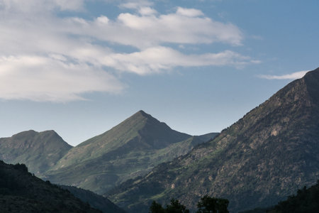 Various mountains in pale green, trees & some foliage under a blue sky with big clouds in Spain