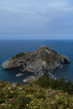 Vertical shot containing some foliage at the front, the whole dragonstone-gaztelugatxe island at the...