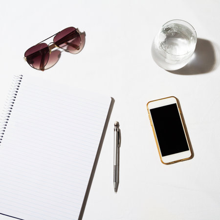 Sunglasses water phone and notepad on a white table from overheadの写真素材