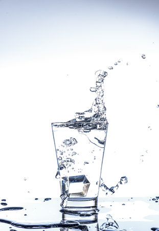 Ice dropping and water splashing in the glass with reflection on the mirror table and isolated on...