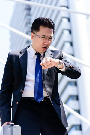 The asian businessman has holding a black bag and looking on watch in hurrying time.の写真素材