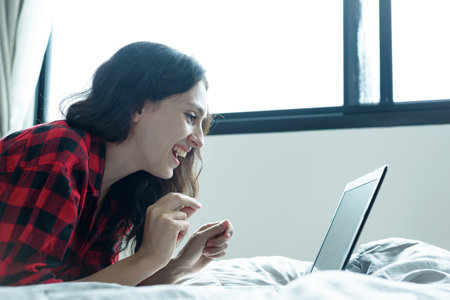 Beautiful woman working on a laptop with smiling and lying down on the bed at a condominium in the morning.の写真素材