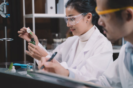 Researcher woman are drop a liquid to a bottle in her hand and researcher man are look to substance and note to notebook in his hand at laboratory.の写真素材