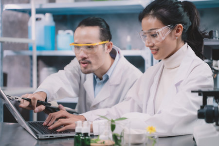 Two research people are comparing information one is working behind a laptop and research man is making notes on a pen and pointing to a laptop in his notebook in a laboratory at a tableの写真素材