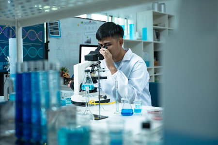 Asian scientist wearing lab coat and gloves using a microscope in a high tech laboratory focused on DNA research biotechnology healthcare and scientific innovationの写真素材