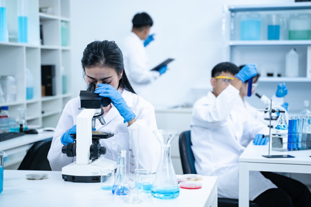 Asian scientist wearing lab coat and gloves using a microscope in a high tech laboratory focused on...