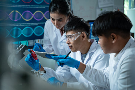 Three young scientists in lab coats working together in a laboratory examining a petri dish for microbiology biology and medical research with focus on education and innovationの写真素材