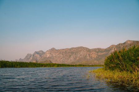 The mountain looms over a green landscape where a swamp meets a tranquil lake, water reflecting the sky, a single cloud drifting in peaceful, untouched nature.の写真素材