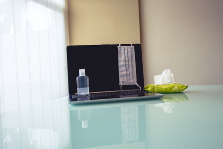 Photo of a laptop with cleaning and health objects such as face mask or disinfectant. Symbolizing new way of working at home.の写真素材
