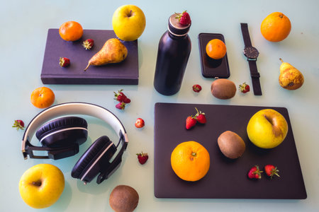 Table with different black objects, laptop, watch, bottle, phone, headphones and a book, organized. And different fruits such as oranges, pears, apples etc decorating it.の写真素材