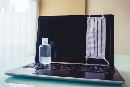 Photo of a laptop with cleaning and health objects such as face mask or disinfectant. Symbolizing new way of working at home.の写真素材
