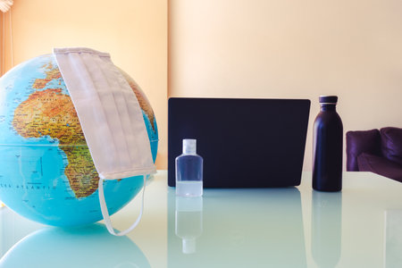 A table on a desk with different objects like a world ball with a face mask, disinfectant, black bottle and a laptop. Office with an international style or virus touch.の写真素材