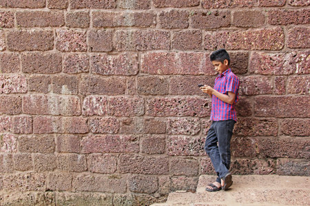 Smart young man working seriously on smartphone, leaning against red brick wallの写真素材