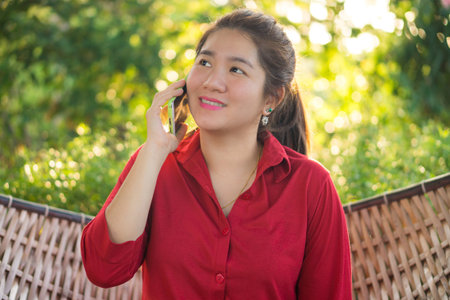 Smiling Thai lady is talking on mobile phone to communication with another people in a wicker bamboo hammockの写真素材