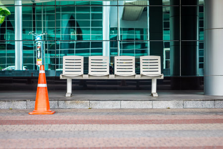 A 4-seat bus waiting bench with a traffic cone at a modern bus stop station. The seats are white and are made of metal.の写真素材