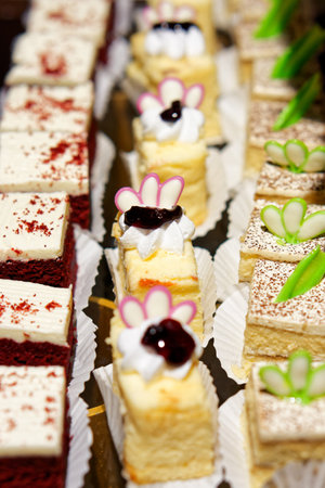 Assorted Mini Cakes with Decorative Toppings. Bite-sized cakes in wrappers, featuring various flavors such as vanilla, chocolate. Each piece is adorned with whipped cream, jam, and edible sugar.の写真素材