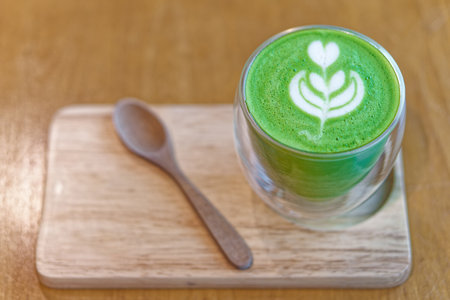Matcha Latte with Beautiful Creamy White Foam Latte Art on a Wooden Tray. A freshly made green matcha latte served in a double walled glass cup, featuring a delicate latte art design on top.の写真素材