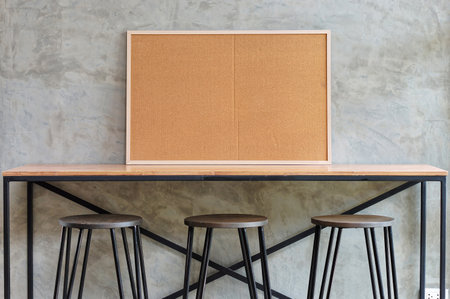 Modern Industrial Workspace with Cork Board and Stools. A contemporary workspace featuring a light wood tabletop, three metal and wood stools, and a cork bulletin board against a textured gray wall.の写真素材