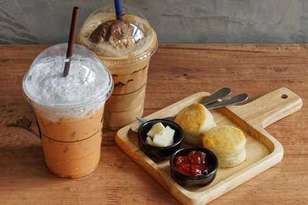 Iced Thai Tea and Coffee with Scones and Jam. A delightful afternoon tea set featuring iced Thai tea and coffee, served with two fluffy scones, butter, and strawberry jam on a wooden tray.の写真素材